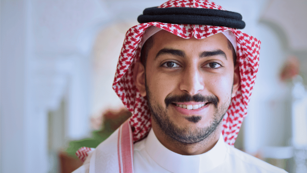 صورة مقربة لشاب سعودي مبتسم يرتدي الزي الرسمي (الثوب والشماغ الأحمر والأبيض والعقال الأسود). تعابير الوجه توحي بالثقة والإيجابية، ويظهر جزء من خلفية داخلية مضيئة وناعمة. يمكن استخدام هذه الصورة لتمثيل العامل السعودي في سياق موضوع حقوق وواجبات العمل.