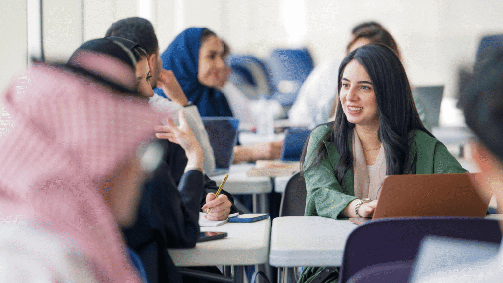 لقطة واسعة لبيئة تعليمية عصرية داخل قاعة محاضرات أو ورشة عمل. تظهر مجموعة متنوعة من الطلاب السعوديين الشباب (طلاب وطالبات سعوديات) منهمكين في نقاش أكاديمي جماعي وتبادل الأفكار. ترتدي إحدى الشابات ذات الشعر الداكن سترة خضراء زيتونية وتستخدم كمبيوتر محمول (لابتوب)، بينما يظهر في المقدمة شاب يرتدي الشماغ والغترة الحمراء والبيضاء (الزي السعودي التقليدي للرجال). تعكس الصورة أجواء التعليم العالي في السعودية، الابتكار التعليمي، والعمل الجماعي في المجتمع الأكاديمي السعودي.