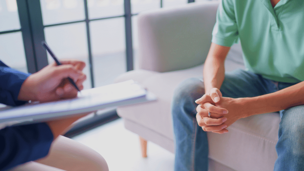 لقطة مقربة لجلسة استشارة نفسية بين أخصائي نفسي (يقوم بتدوين الملاحظات) وشخص مستفيد، تعكس المشهد العملي لـ علم النفس العيادي (الإكلينيكي) والإرشاد النفسي. توضح الصورة الدور المحوري للأخصائيين في تشخيص وعلاج الاضطرابات النفسية، وهي وظائف رئيسية مطلوبة في القطاع الصحي بالمملكة، مما يبرز الفرص الوظيفية في القطاع الصحي والعيادات الخاصة لخريجي علم النفس.