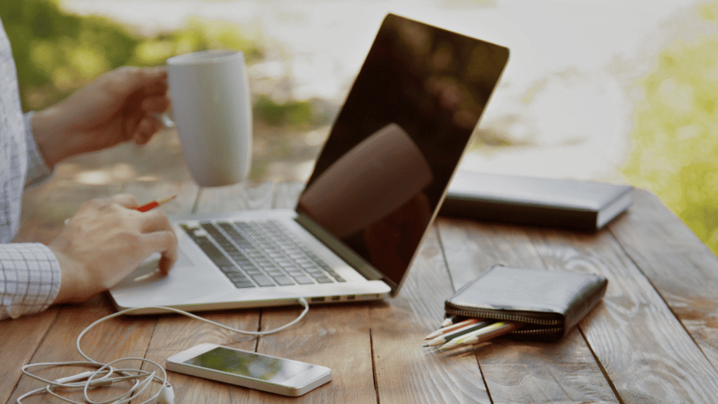 لقطة مقربة تظهر شخصًا يعمل على حاسوب محمول (لابتوب) موضوع على طاولة خشبية في الهواء الطلق أو في حديقة (تظهر خلفية خضراء ضبابية). يمسك الشخص بيده كوبًا أبيض (قد يكون قهوة أو شاي)، وفي يده الأخرى قلمًا أحمر. يظهر على الطاولة هاتف ذكي، وسماعات أذن ملفوفة، ومحفظة جلدية تحتوي على أقلام ملونة. تجسد هذه الصورة مفهوم العمل المرن والعمل من أي مكان (Work From Anywhere)، وتوضح التوازن بين العمل والحياة الشخصية والطبيعة.