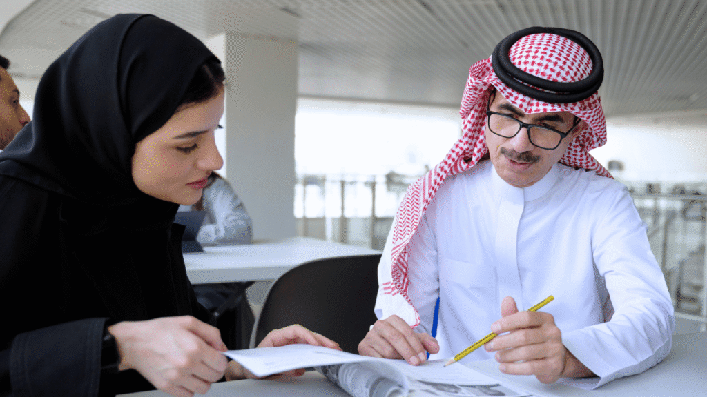 اجتماع عمل احترافي يجمع رجل أعمال سعودي يرتدي الثوب الأبيض والشماغ التقليدي ونظارات طبية، مع سيدة أعمال سعودية ترتدي العباءة والحجاب الأسود الأنيق. يظهر الرجل وهو يشير بقلم رصاص إلى وثيقة أو جريدة مالية، مما يوحي بـتحليل البيانات أو استراتيجيات العمل أو التدريب المهني. تعكس الصورة الشراكة المهنية والقيادة السعودية في بيئة العمل، وتبرز دور المرأة السعودية في الأعمال والتمكين الاقتصادي.