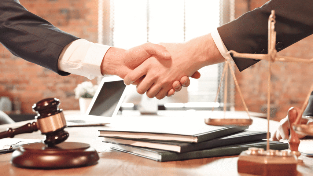 مصافحة بين شخصين يرتديان بدلات رسمية في مكتب محاماة مع ظهور ميزان العدالة ومطرقة القاضي على الطاولة.