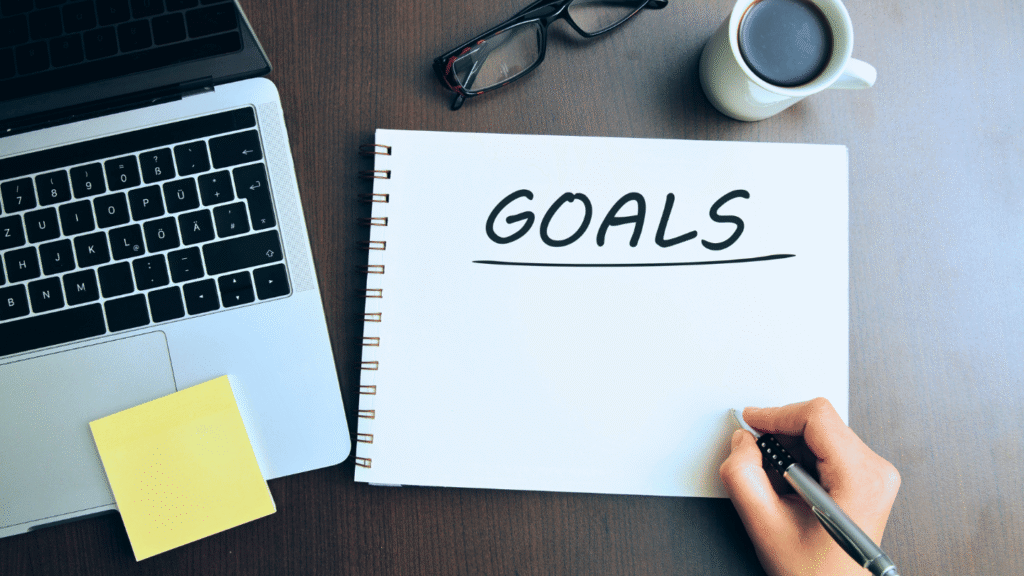 صورة من الأعلى لشخص يكتب كلمة GOALS (أهداف) في دفتر ملاحظات، يمثل صياغة هدف وظيفي جاهز لحديثي التخرج.
