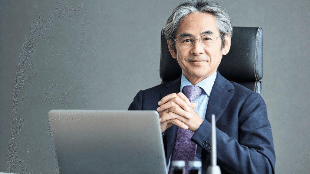 مدير تنفيذي أو مسؤول كبير (من أصل آسيوي) ذو شعر رمادي يجلس خلف مكتبه وأمامه جهاز حاسوب محمول، يمثل القيادة والخبرة في مجال إدارة الأعمال.