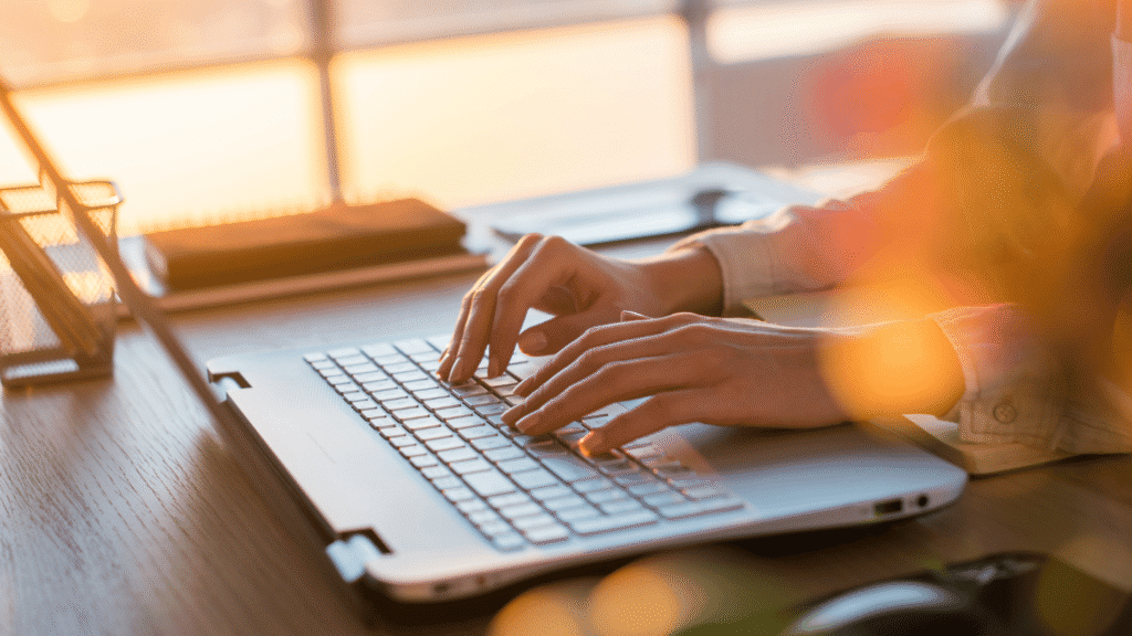 لقطة مقربة تظهر يدي شخص يكتبان على لوحة مفاتيح حاسوب محمول  (لابتوب) يشتغل في مجال العمل الحر. تظهر الإضاءة الطبيعية الدافئة (ربما ضوء الشمس وقت الشروق/الغروب) تدخل من النافذة في الخلفية، مما يخلق انعكاساً لطيفاً على الشاشة وعلى سطح المكتب الخشبي. يظهر جزء من طاولة عمل مع بعض الأغراض المكتبية.