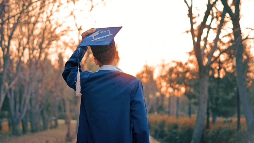 شاب يرتدي زي التخرج الأزرق ويقف في الهواء الطلق عند غروب الشمس بينما يضع يده على قبعة التخرج.