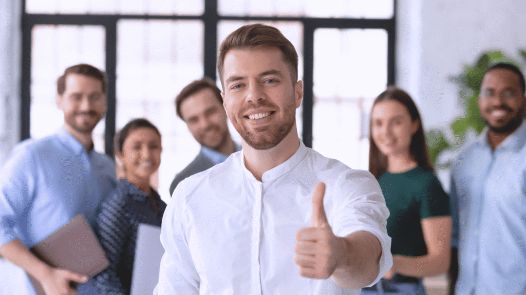 فريق عمل متعدّد الأعراق يقف بابتسامة وثقة، يظهر فيه رجل في المقدمة يرتدي قميصاً أبيض ويشير بإبهامه للأعلى، يمثل بيئة عمل صحية وإيجابية بعد التعافي من الإجهاد.