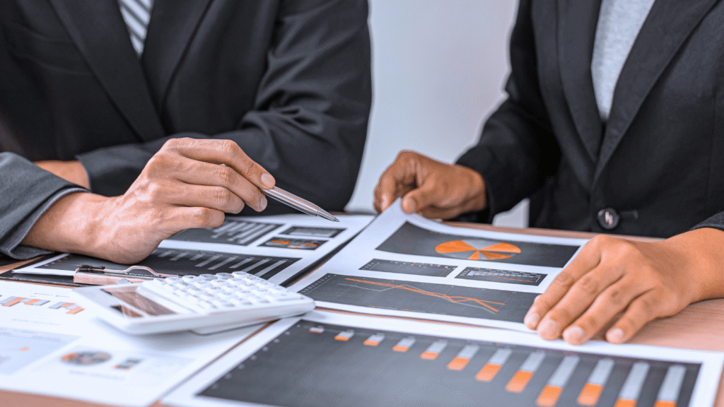 شخصان يرتديان ملابس رسمية (بدلة) ويناقشان تقارير عمل ورسومات بيانية مع وجود آلة حاسبة، تمثل العمل المشترك والمشاورات المالية.