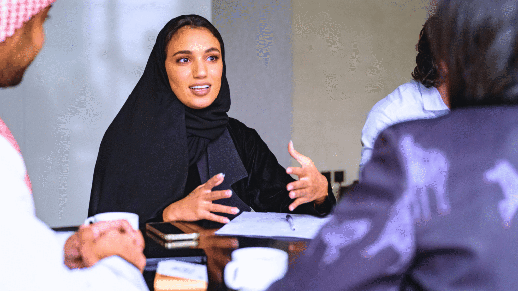 سيدة أعمال سعودية في اجتماع تنفيذي أو جلسة حوارية مهمة. ترتدي الحجاب والعباءة السوداء بأناقة وتتحدث بـانفعال وثقة عالية، مستخدمة إيماءات يدوية معبرة أثناء تقديم عرض أو إبداء رأي استراتيجي. يجلس حولها زملاء في العمل (يظهر جزء منهم بالزي التقليدي). تُبرز هذه اللقطة القيادة النسائية السعودية، مهارات العرض والإلقاء، والثقة بالنفس في سياق البيئة المهنية السعودية.