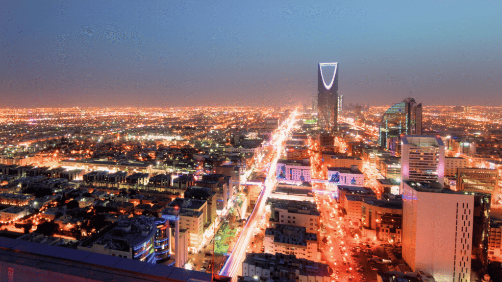 منظر بانورامي ليلي مذهل لـأفق مدينة الرياض، عاصمة المملكة العربية السعودية. يسيطر على المشهد مركز المملكة (Kingdom Centre) الشهير بقمته المضيئة ذات الشكل الماسي (على شكل قبة) والبرج الذي يشبه فتاحة الزجاجات، وهو أيقونة معمارية سعودية. تظهر أضواء المدينة الساطعة وحركة السيارات الطويلة المتمثلة في خطوط ضوئية على طول الشوارع، مما يعكس التطور العمراني السعودي، المدينة الذكية، وديناميكية الحياة الليلية في الرياض.