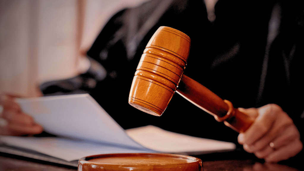 صورة مقربة لمطرقة القاضي الخشبية أثناء جلسة محاكمة، مع ظهور يد القاضي وهو يراجع المستندات. ترمز الصورة إلى إصدار الأحكام القضائية، سلطة القاضي، إجراءات التقاضي، وقرارات المحكمة. مناسبة للمقالات المتعلقة بالقانون، حقوق المتقاضين، القضايا المدنية والجنائية، وآليات القضاء السعودي.